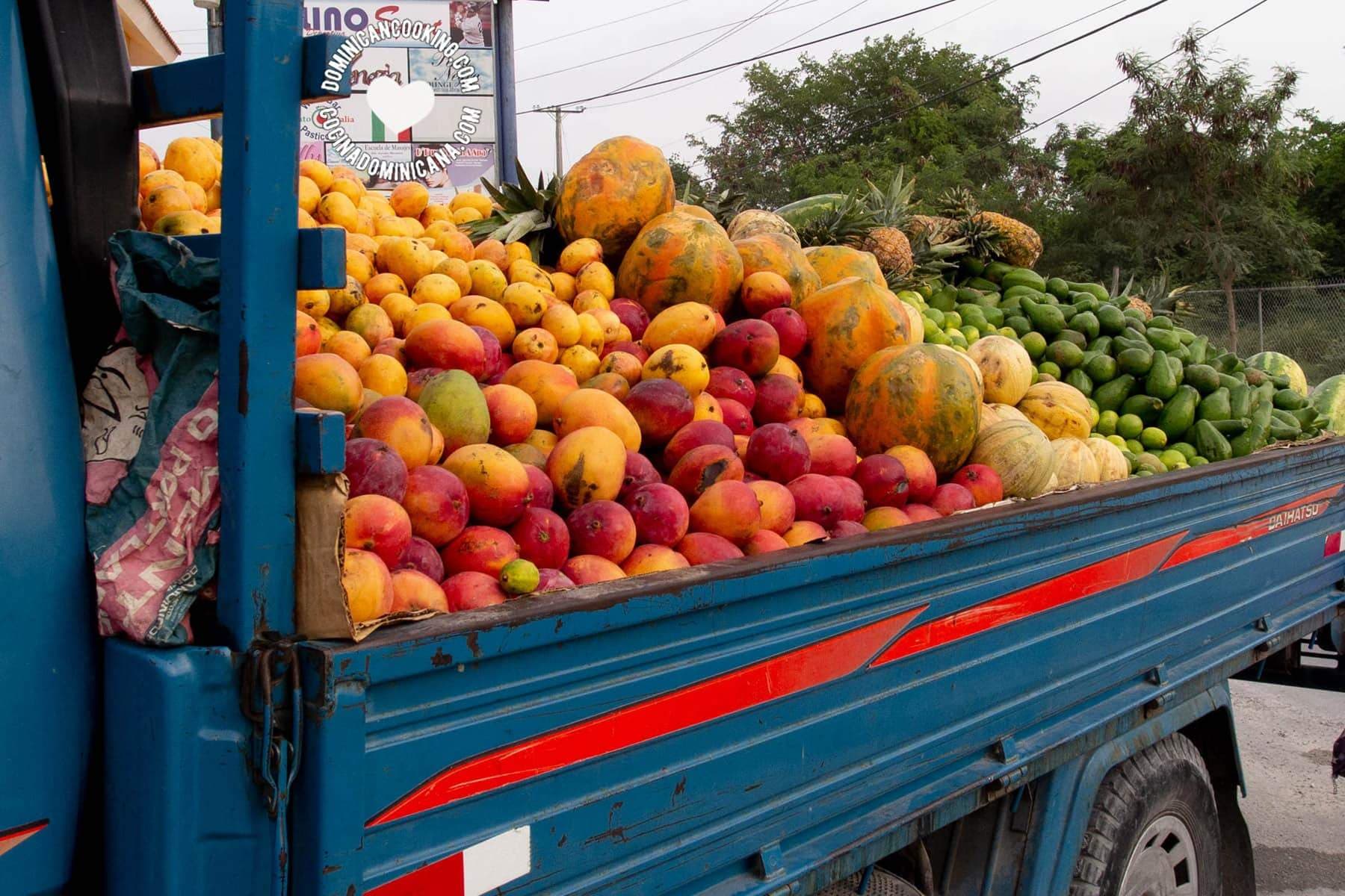 A blue truck bed filled with a colorful assortment of tropical fruits, including mangoes, papayas, pineapples, and limes, typical of Dominica's local markets.