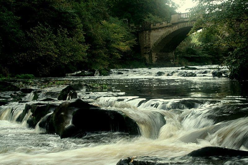 River Almond weir with rushing water over rocks, stone bridge, and lush green woodland in summer.
