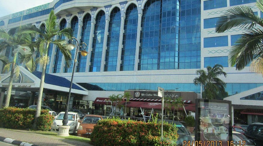 Modern commercial building with glass facade and arched windows, palm trees, parked cars, and street-level shops in the Bandar Seri Begawan city center.