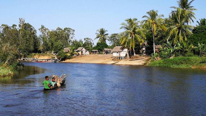 Two boys transport a bundle of wood across a wide river near a sandy bank lined with palm trees and stilt houses.