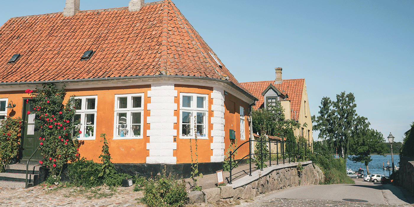 A sunny streetscape in Præstø featuring a vibrant orange building with white quoins and a red-tiled roof, situated near a cobblestone path and waterfront.