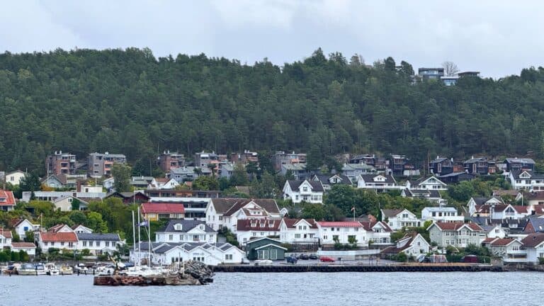 A scenic waterfront view of Drøbak featuring a dense cluster of houses on a forested hillside, with boats docked in the harbor below.
