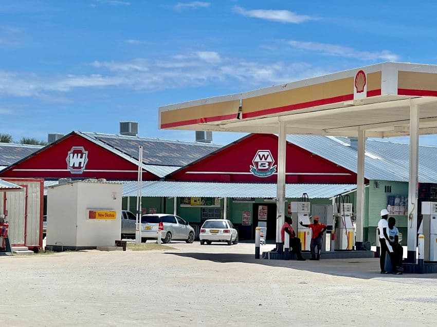 Exterior of a Shell fuel station in Okahao, Ongandjera, with red-roofed buildings, parked cars, and workers at the pumps under a clear blue sky.