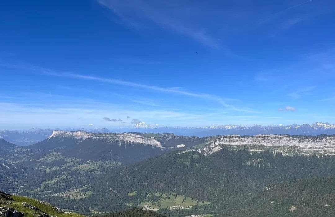 Wide landscape photograph showing forested mountain ridges, rocky limestone cliffs, and distant snow-capped peaks under a clear blue sky.