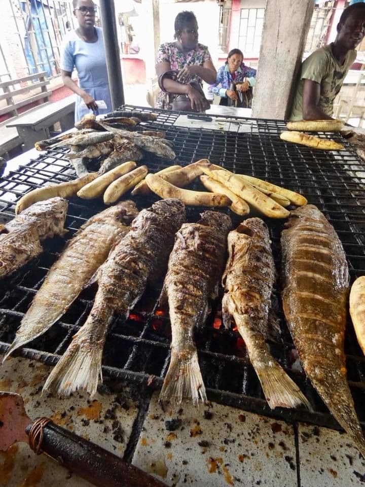 Whole fish and plantains grilling on a large outdoor grill at the Limbe beach market, with vendors and customers in the background