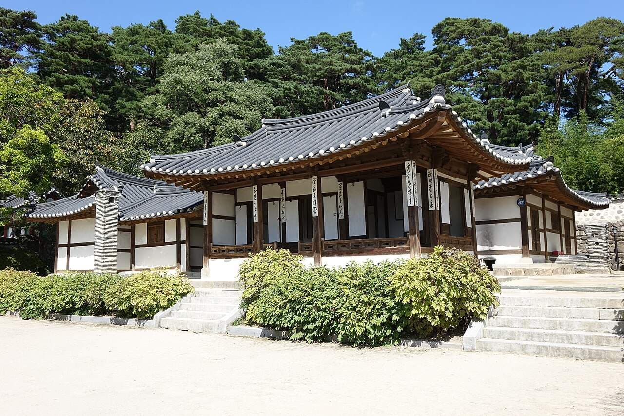 Traditional Korean hanok exterior with tiled roofs, white walls, and calligraphy scrolls, surrounded by greenery.