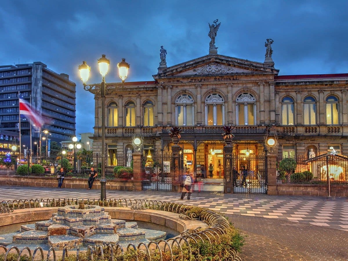 Teatro Nacional facade at dusk in San José's central plaza, with fountain, lampposts, and a few pedestrians.