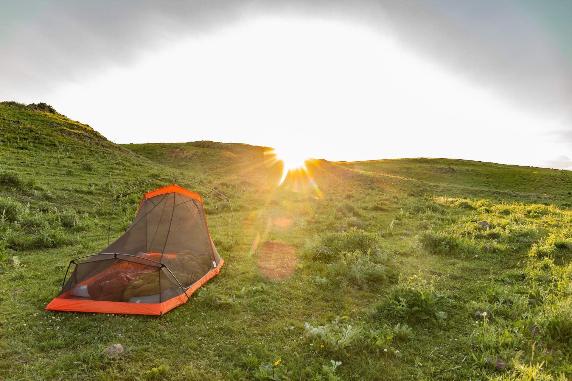 Orange camping tent on a grassy hillside with rolling green hills and a bright sun flare on the horizon.