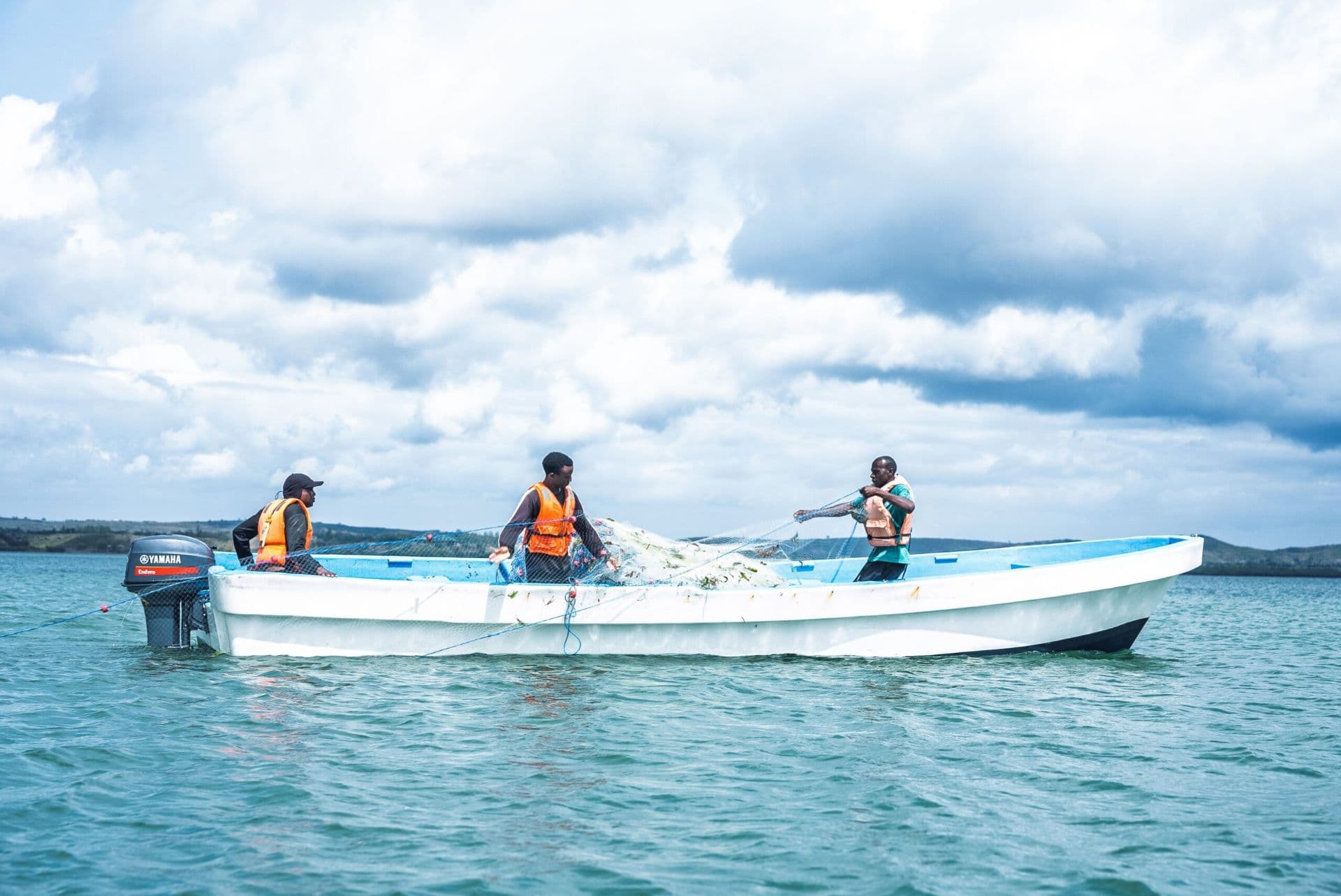 Three men in life vests handle fishing nets aboard a white motorboat on calm turquoise water near the Tanzania coast.