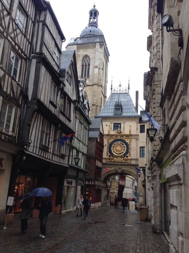 Cobblestone street in Roncherolles-sur-le-Vivier with half-timbered buildings, a clock tower, and the church steeple in the background