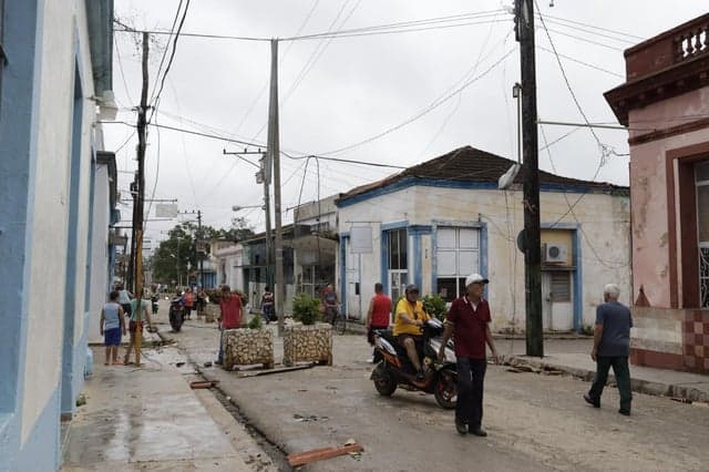 A quiet street scene in San Antonio de los Baños featuring colonial-style buildings, pedestrians, and a motorcyclist navigating the road.