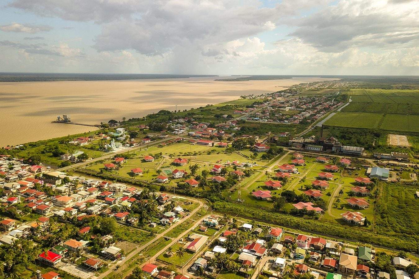 Aerial view of Skeldon, a riverside town on the Corentyne River estuary in Guyana, with red-roofed houses, green fields, and a wide muddy river stretching to the horizon.