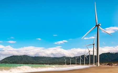 A scenic coastal view featuring a sandy beach, turquoise ocean waves, and a row of wind turbines against a backdrop of mountains and blue sky.