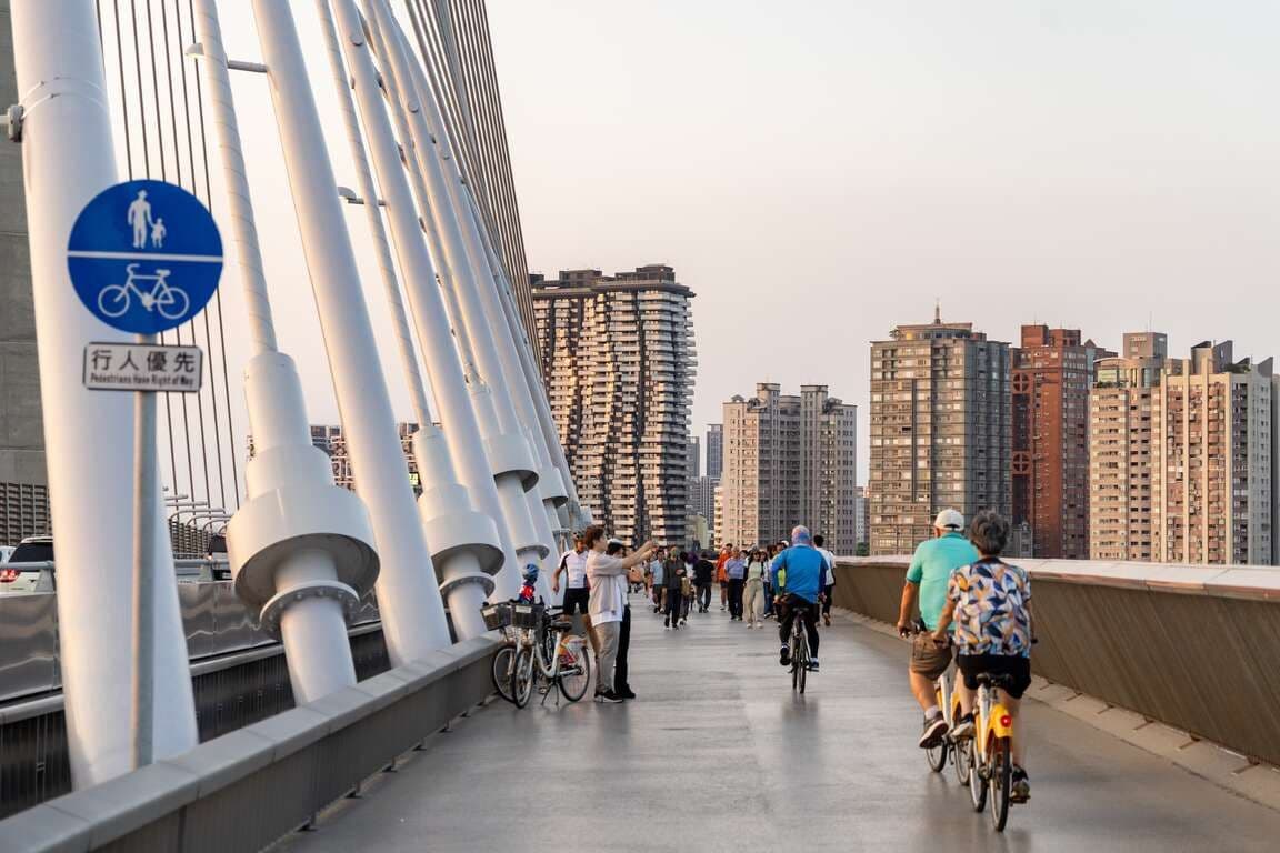 Cyclists and pedestrians use a dedicated path on a modern cable-stayed bridge in an urban setting, featuring a blue sign indicating pedestrian priority.