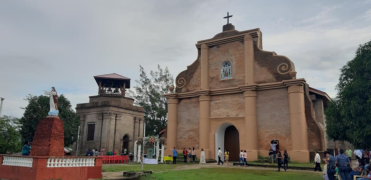 Exterior view of a historic brick church in Piddig featuring a bell tower, statue, and visitors gathered on the grounds.