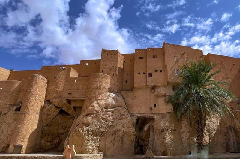 Wide view of earthen kasbah architecture in Smala, M'zab Valley, with a palm tree and rocky terrain under a blue sky.