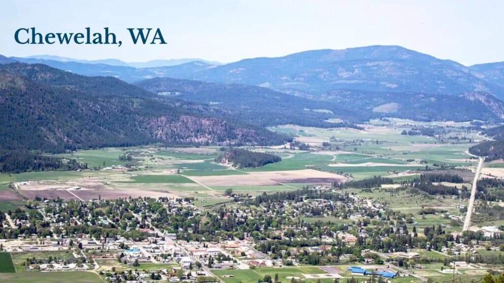 Aerial view of Chewelah, WA, showing a town nestled in a valley surrounded by green fields and forested mountains under a clear sky.