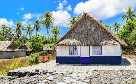 A thatched-roof hut with white and blue walls on a rocky shoreline backed by coconut palms under a bright blue sky, near Ambo Village, Tarawa Atoll, Kiribati.