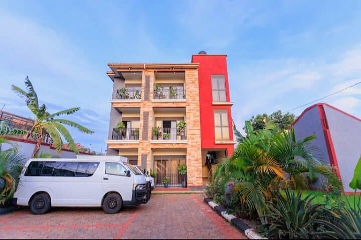 Exterior view of a multi-story guesthouse with red and beige facade, featuring balconies with plants and a white van parked on a paved driveway.