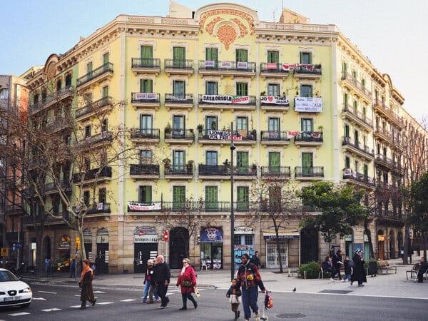 Street-level view of a yellow corner building with balconies, banners, and pedestrians crossing the plaza in front