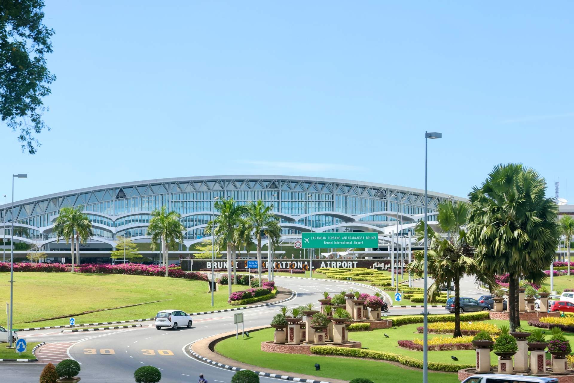 Exterior view of Brunei International Airport terminal with curved glass facade, landscaped grounds, palm trees, and signage.