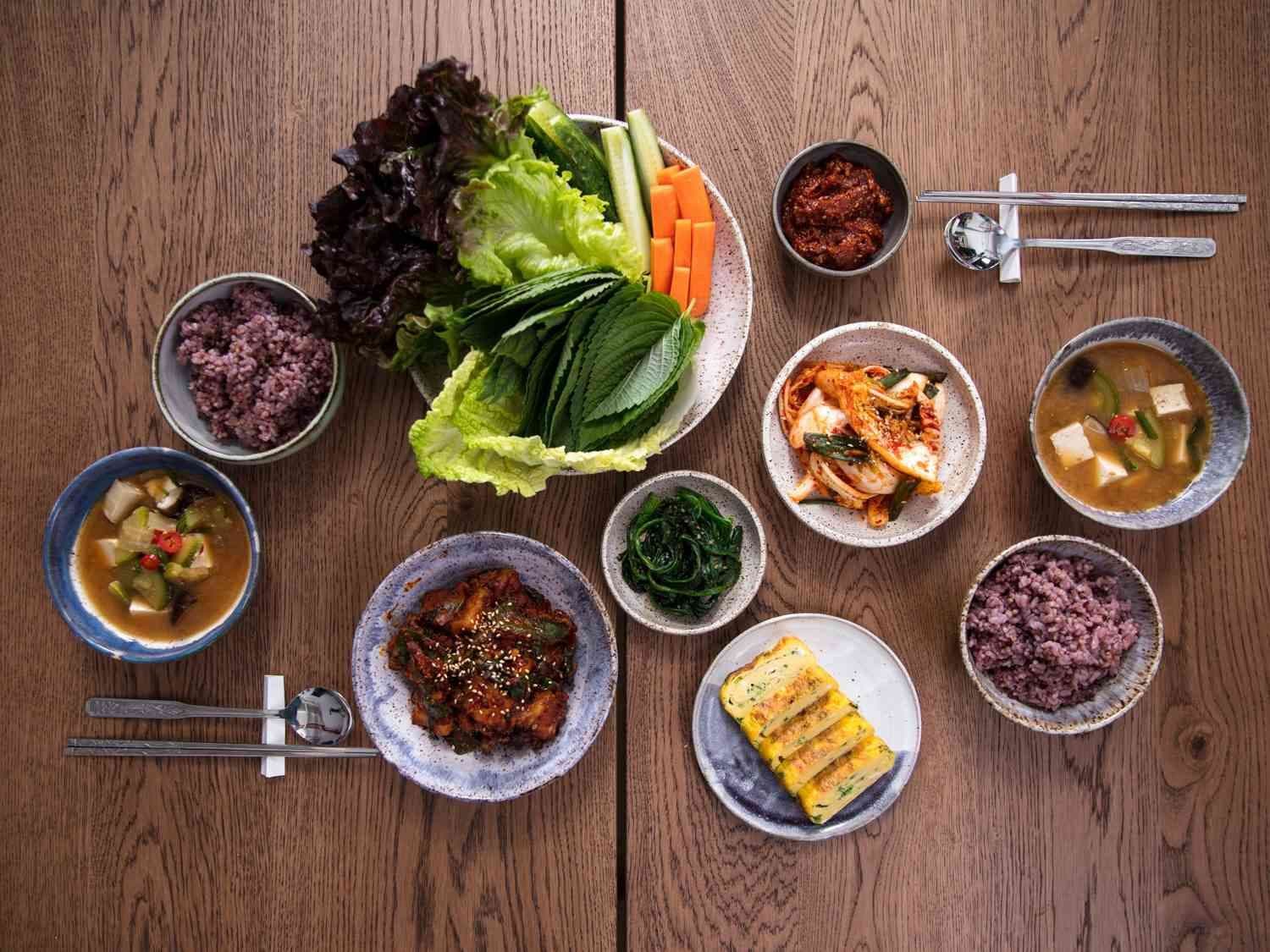 Overhead shot of a Korean-style communal table with rice, soups, banchan, lettuce, and metal cutlery on wood.