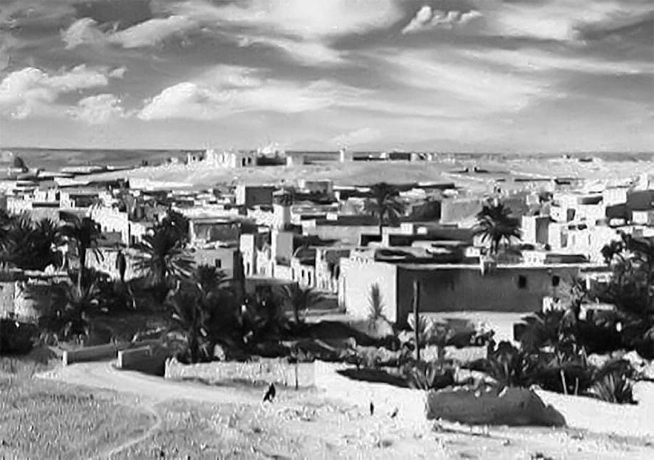 Black-and-white photograph of a traditional stone village with flat-roofed houses, palm trees, and a wide streetscape under a cloudy sky