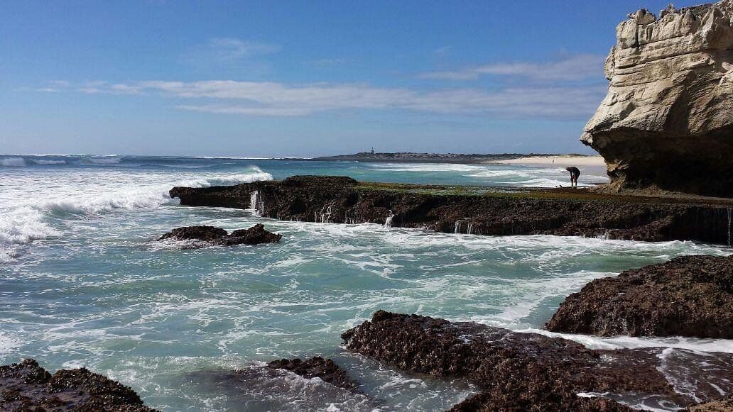 Coastal scene with turquoise surf, dark rocky outcrops, and a large white cliff on the right under a blue sky.