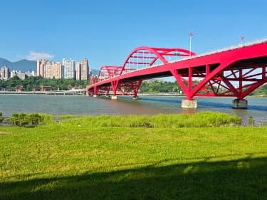 Red arched bridge over a river with green grassy bank, distant high-rises and mountains under a clear blue sky.