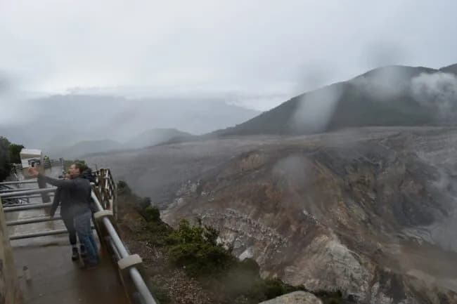 A person stands at a railing overlooking a misty volcanic crater landscape with mountains in the background.