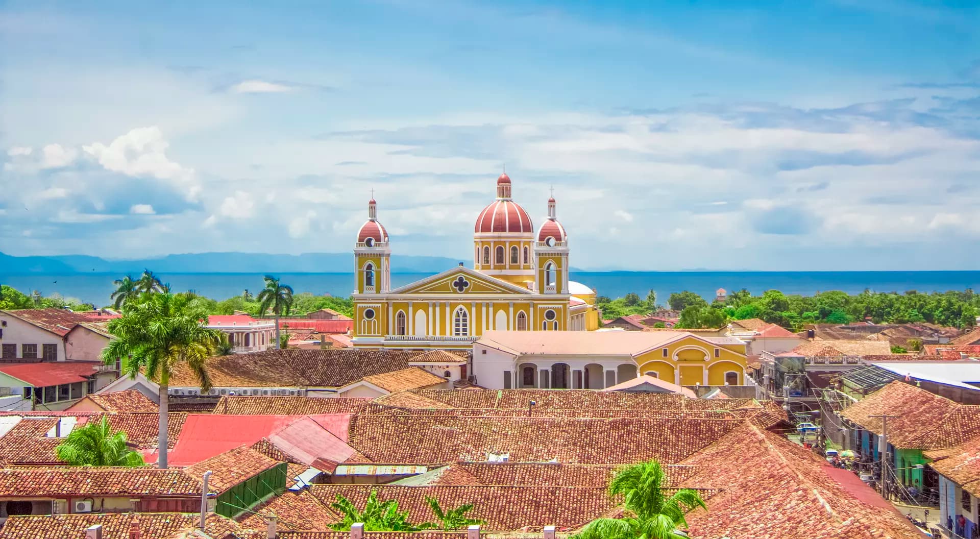 Aerial view of a yellow colonial church with red domes rising above tiled rooftops in a coastal town.