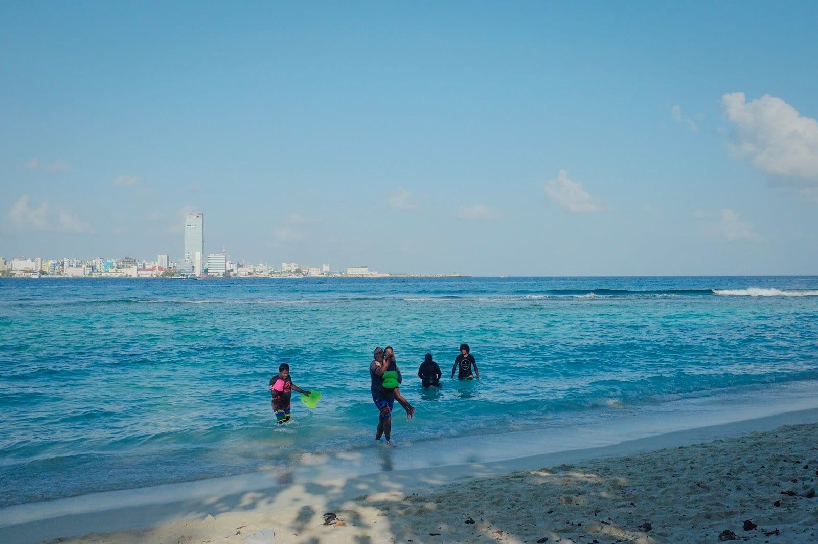Wide shot of Villingili Public Beach with turquoise water and Malé skyline in the distance.