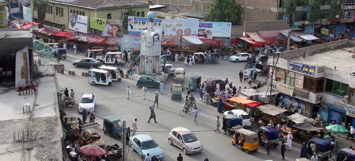 Aerial view of a busy street market in Jalalabad with vehicles, pedestrians, stalls, and surrounding buildings.