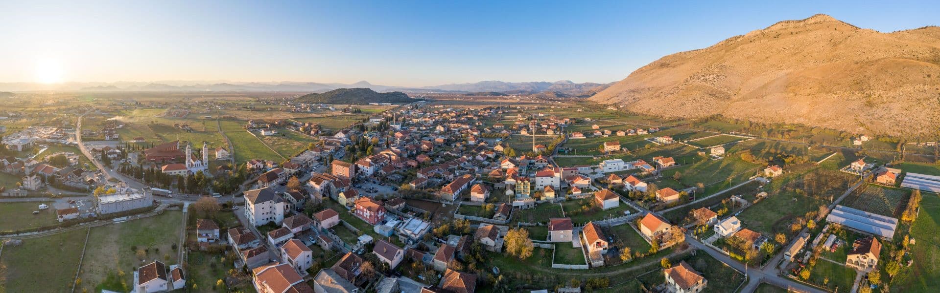 Aerial panorama of Tuzi hillside town with red-roofed houses, green fields, and surrounding mountains.