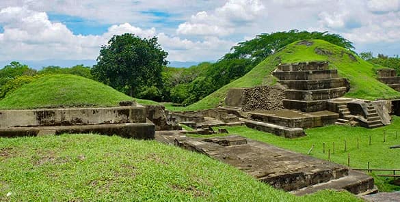 Grass-covered mounds and stone ruins under a partly cloudy sky, with trees in the background