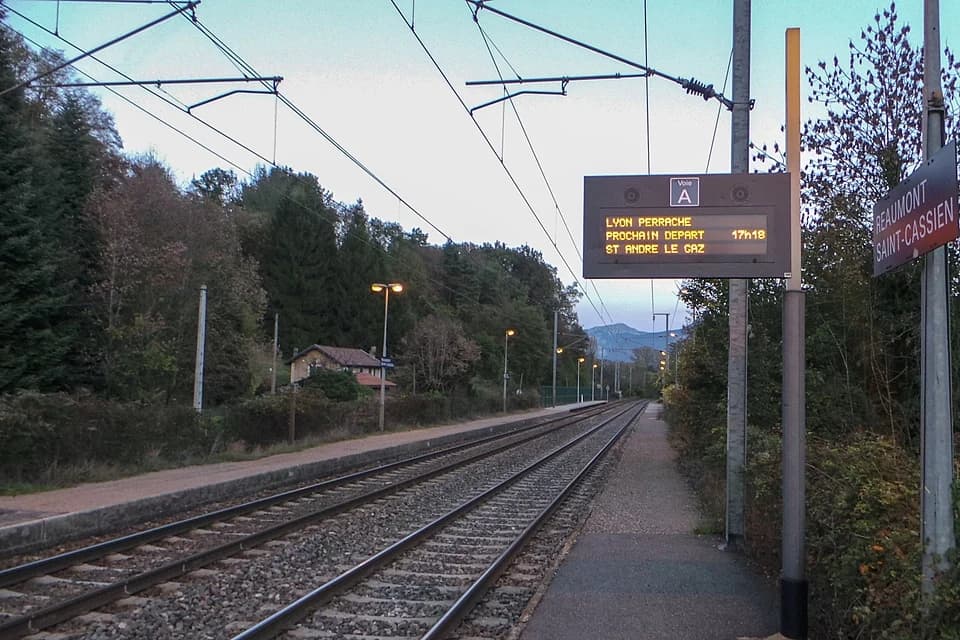 Small rural SNCF station platform with tracks, overhead wires, and a digital departure board reading Saint-Cassien.
