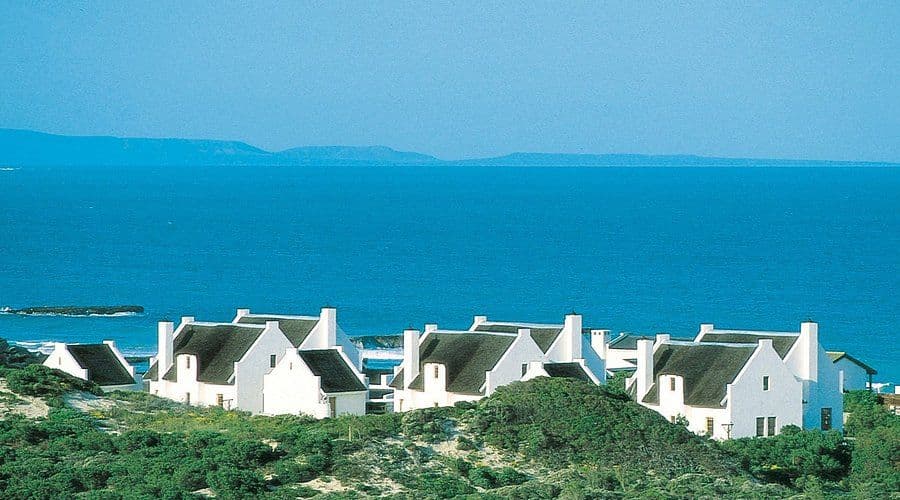 Aerial view of white thatched-roof buildings on a coastal hillside overlooking the blue ocean and distant mountains.