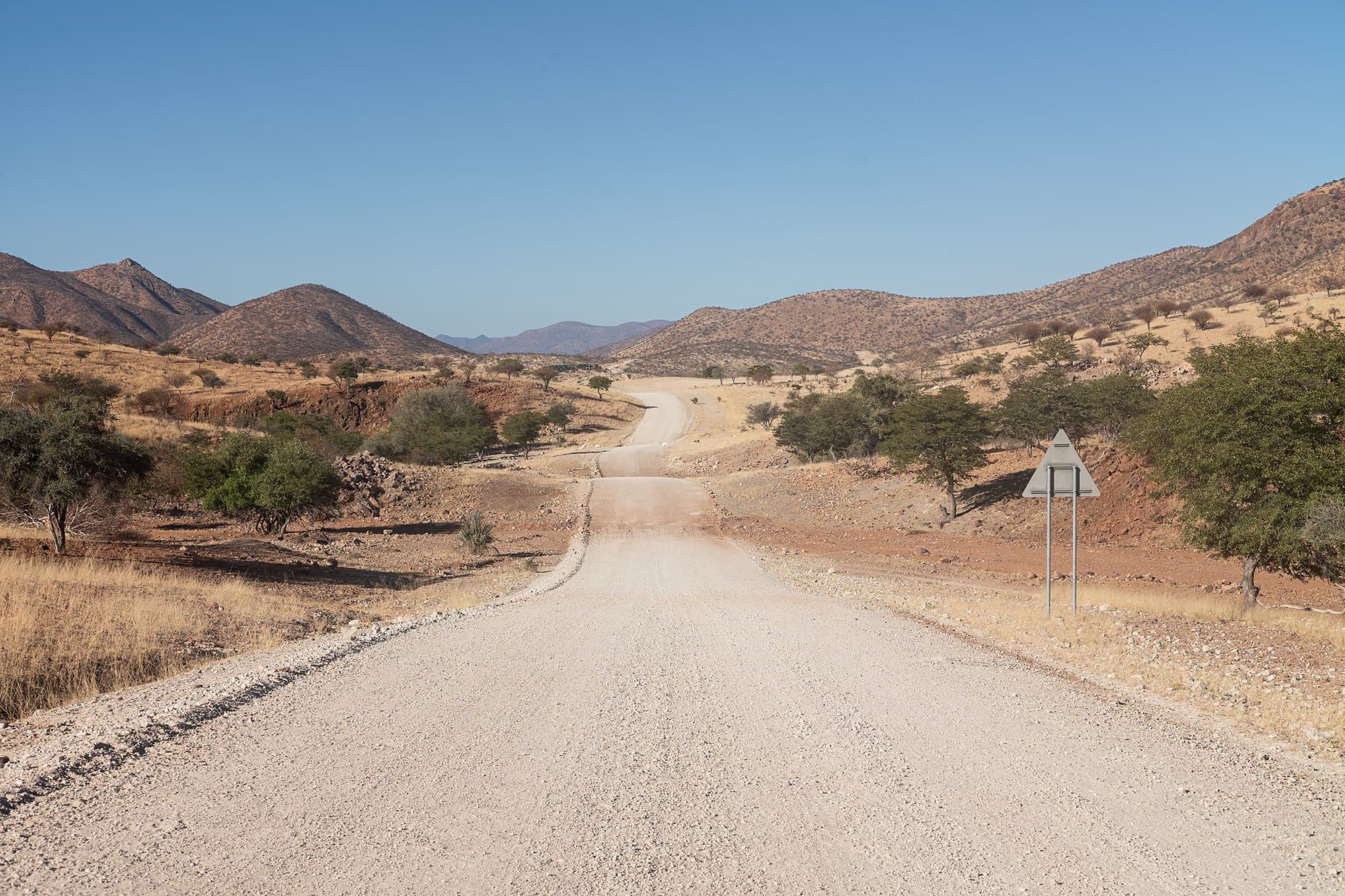 A gravel road winds through an arid, mountainous landscape with scattered trees and a clear blue sky, typical of the Namibian outback.