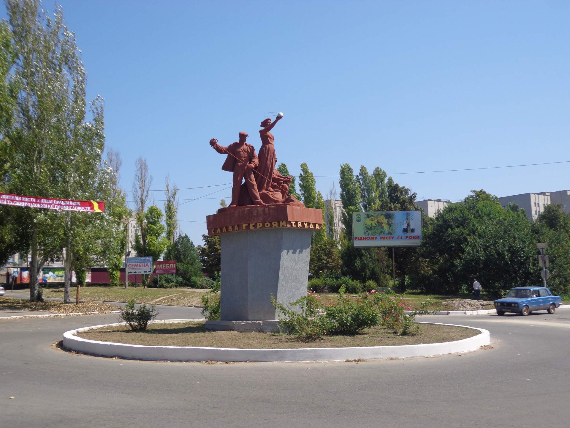 Soviet-style statue of a worker on a concrete pedestal in a Dniprorudne town roundabout with trees, signage, and a blue car.