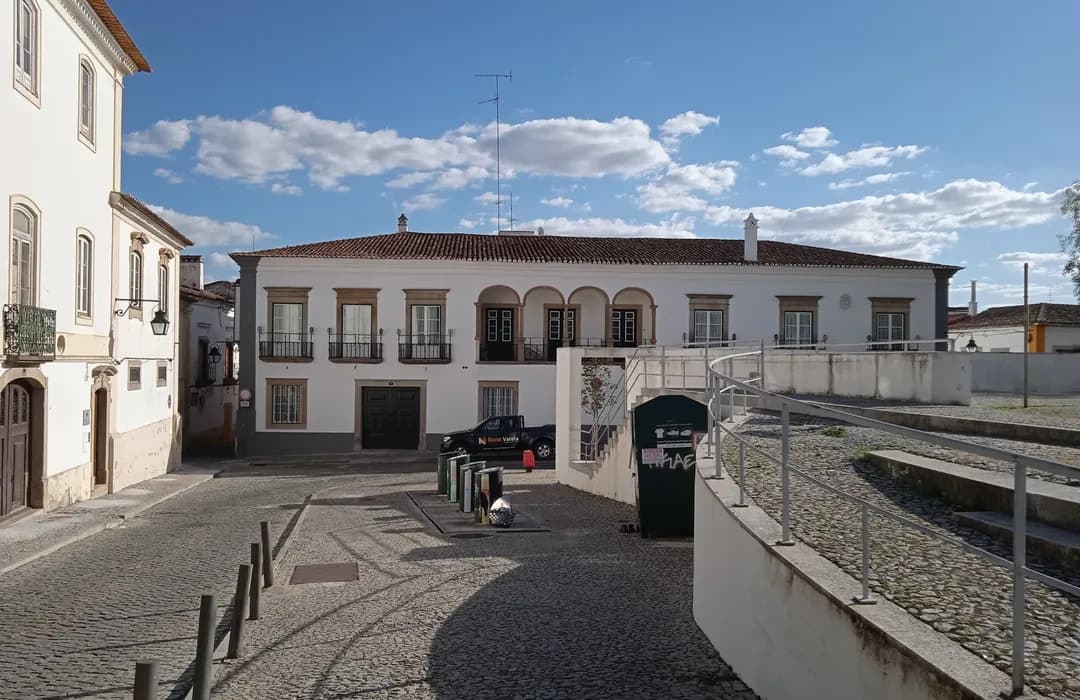 Sunlit cobblestone square in Vimioso featuring traditional white buildings with tiled roofs, arched windows, and a clear blue sky.