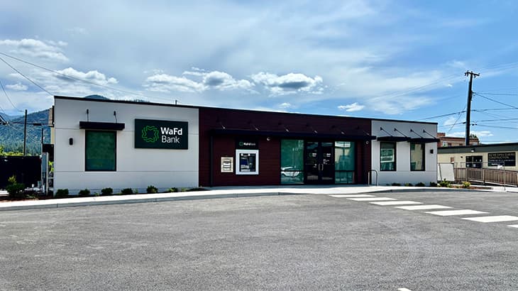Exterior view of a WaFd Bank branch in Chewelah, featuring a white and maroon building facade, parking lot, and mountains in the background.