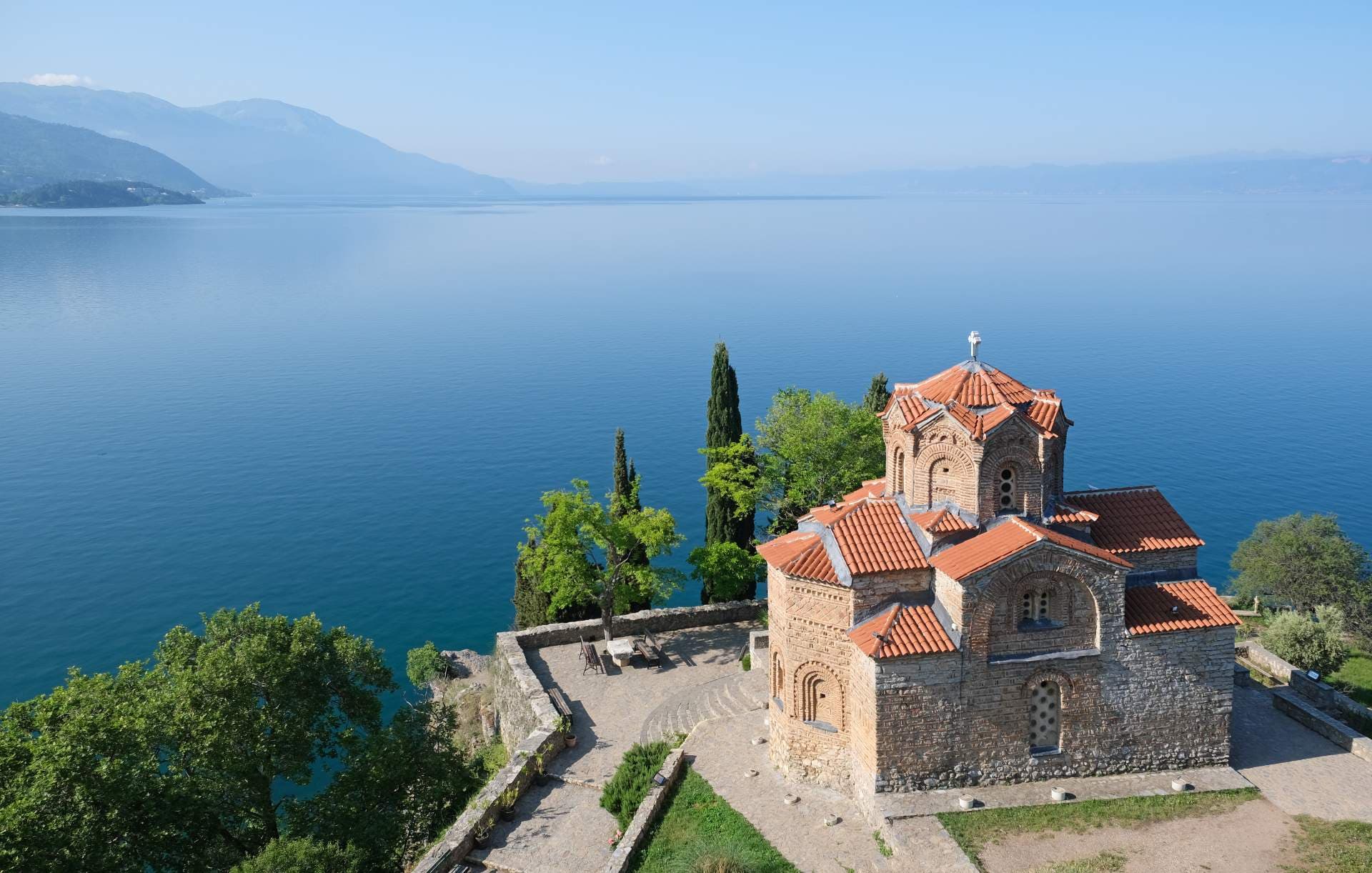 Aerial view of a stone church with red-tiled roofs on a cliff overlooking a large lake and distant mountains.
