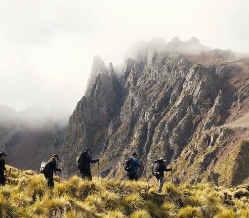 Hikers traverse a grassy ridge with misty mountain peaks in the background, illustrating the elevated atmospheric conditions of the Dubreuil plateau, Mauritius.