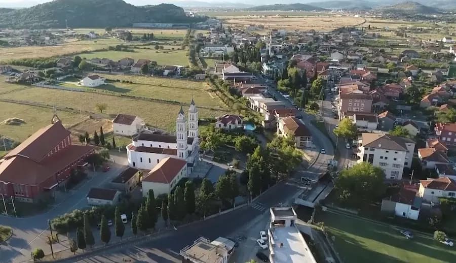 Aerial view of Tuzi with a prominent white church, red-roofed buildings, and surrounding green highlands.