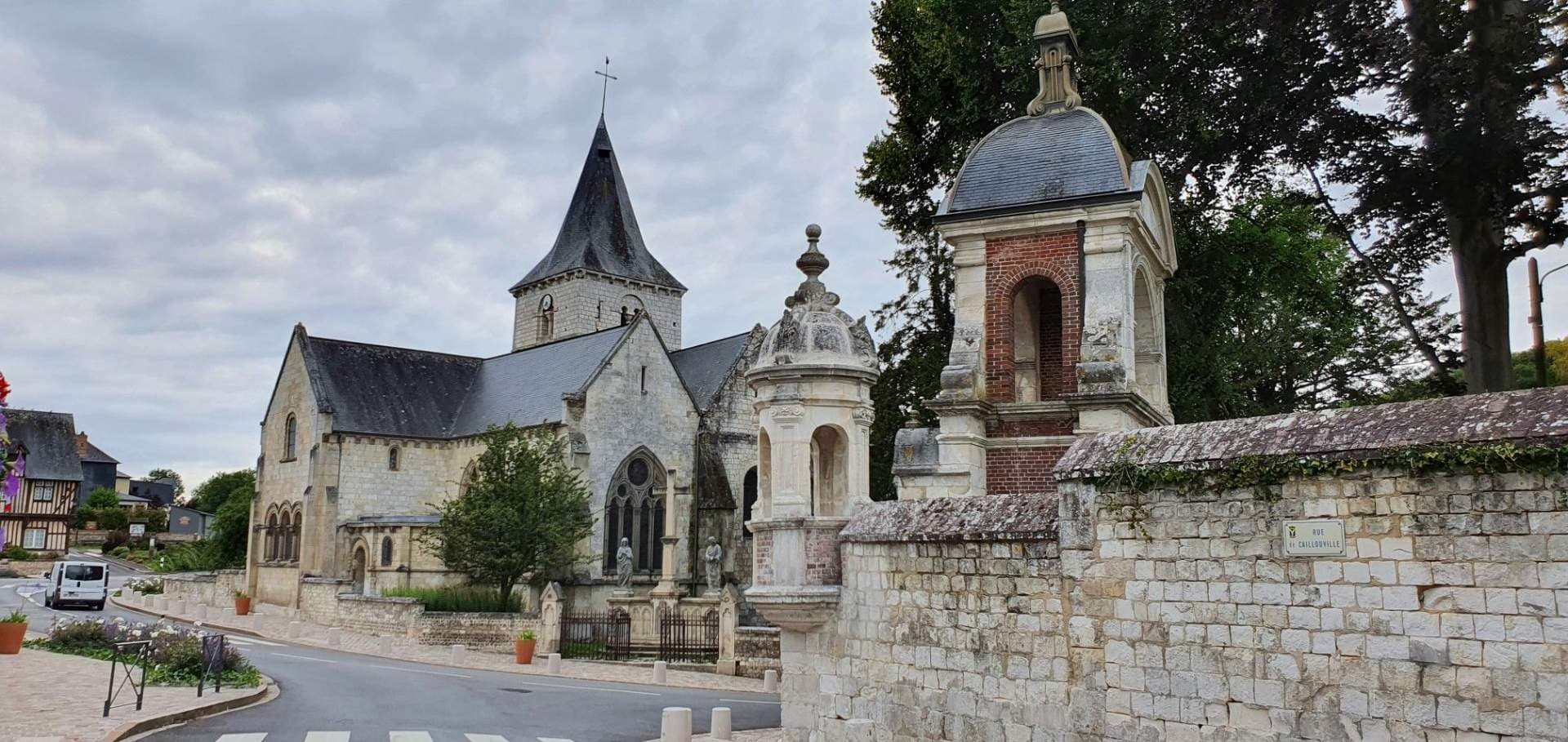 Stone church with spire, ornate chapels, and stone wall in a quiet village under overcast sky near Roncherolles-sur-le-Vivier, France