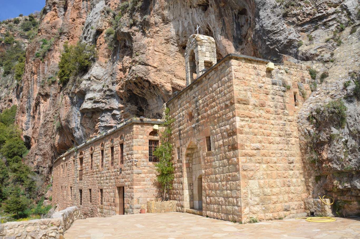 Stone monastery exterior built into a rocky cliff face, with a bell tower, arched entrance, and paved courtyard.