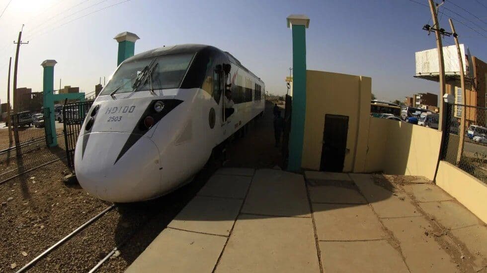 White HD100 2503 train at a small station platform with teal pillars, yellow building, and urban background.
