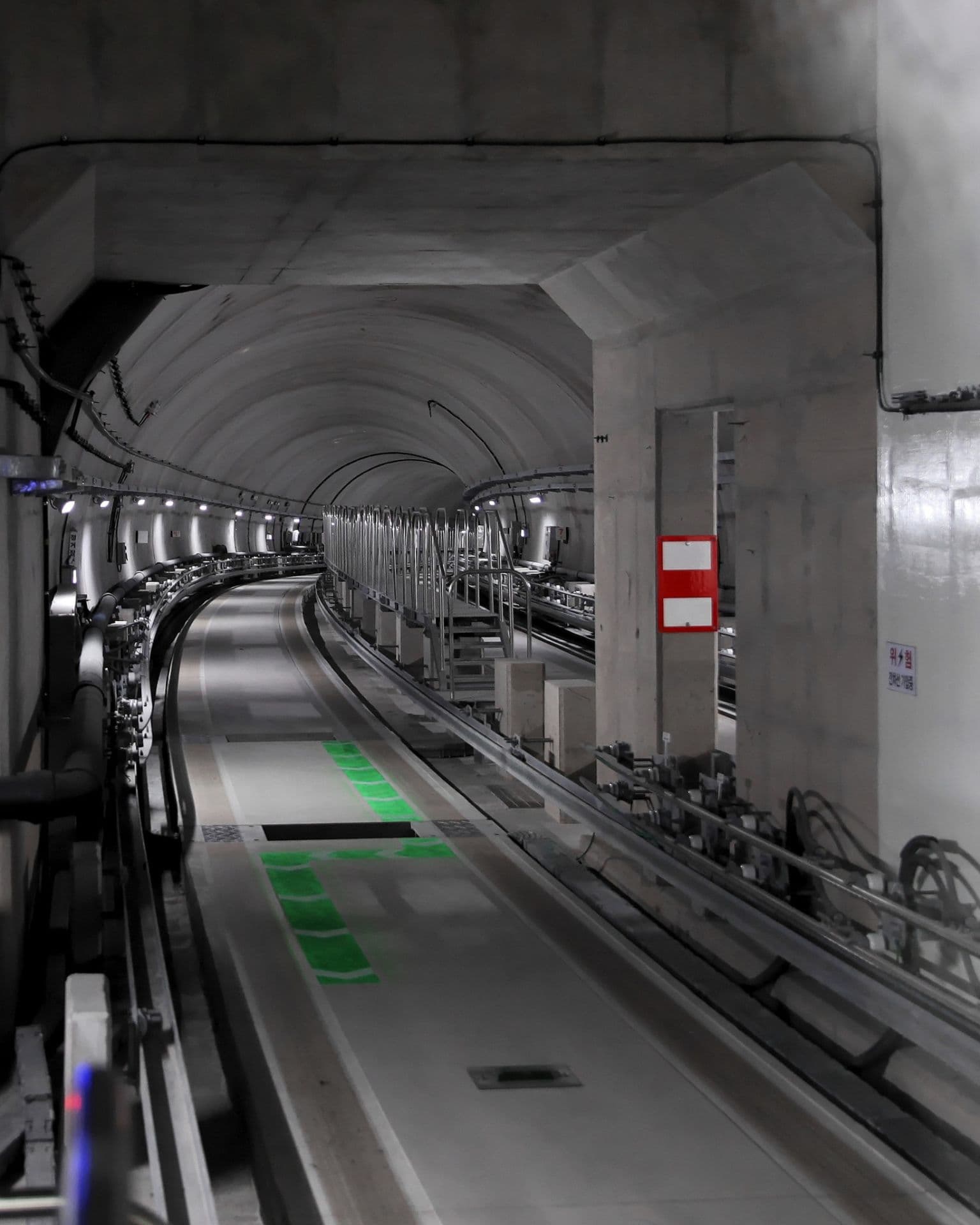 Underground subway tunnel with curved tracks, concrete arch ceiling, green floor markings, and a red sign on a pillar.