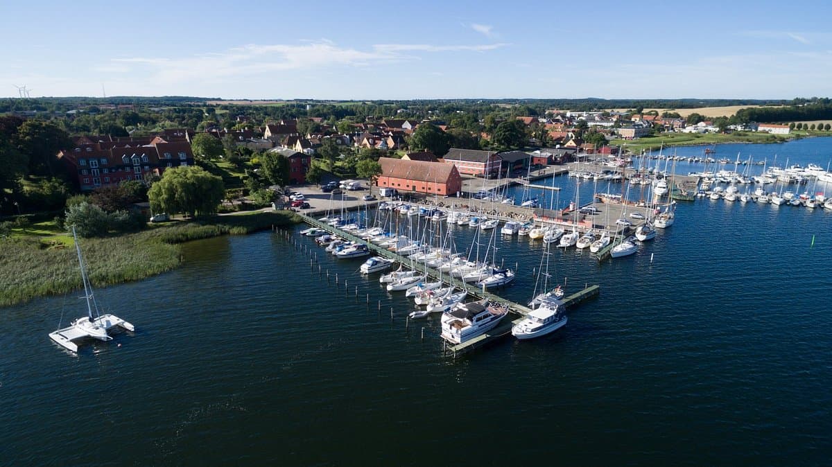 Aerial view of Præstø harbor featuring numerous sailboats docked at piers, a red brick building, and a coastal town under a blue sky.