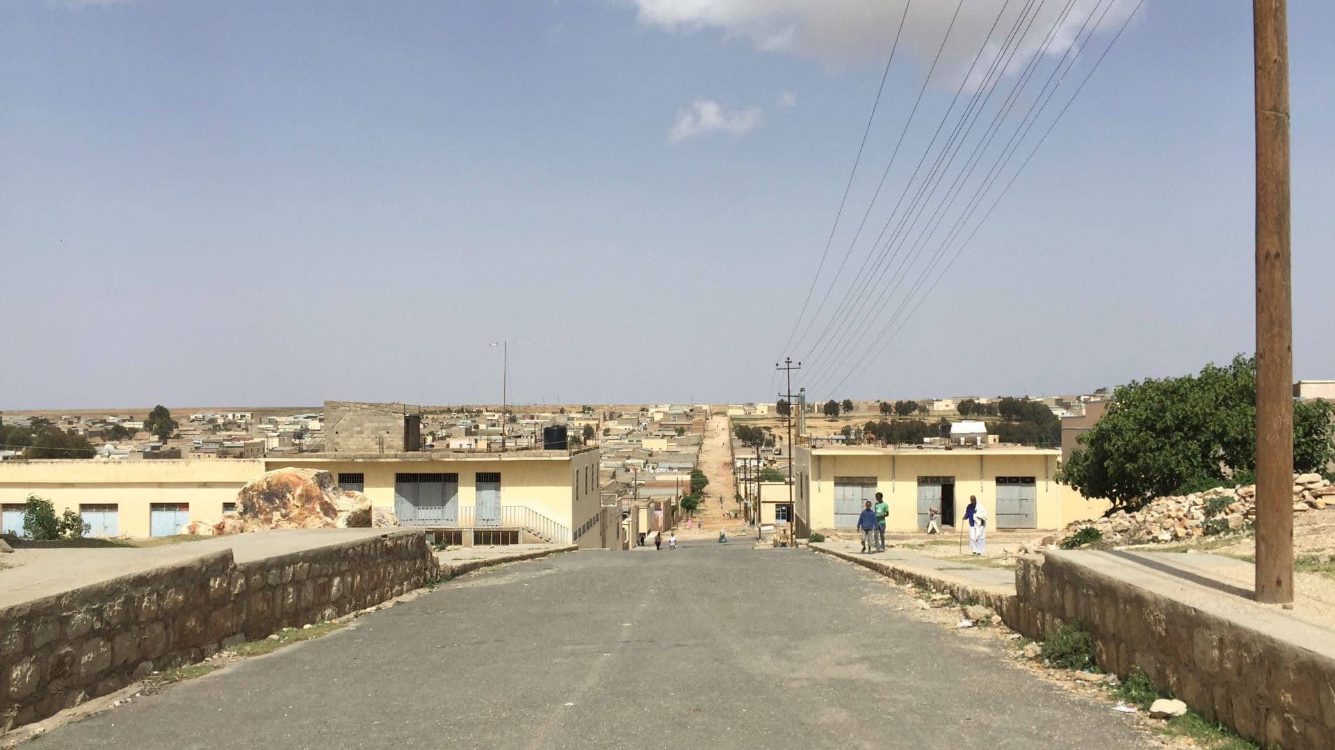 Quiet paved street in Adi Keyh, Eritrea, with low yellow buildings, stone walls, power lines, and a few distant pedestrians under a hazy sky.